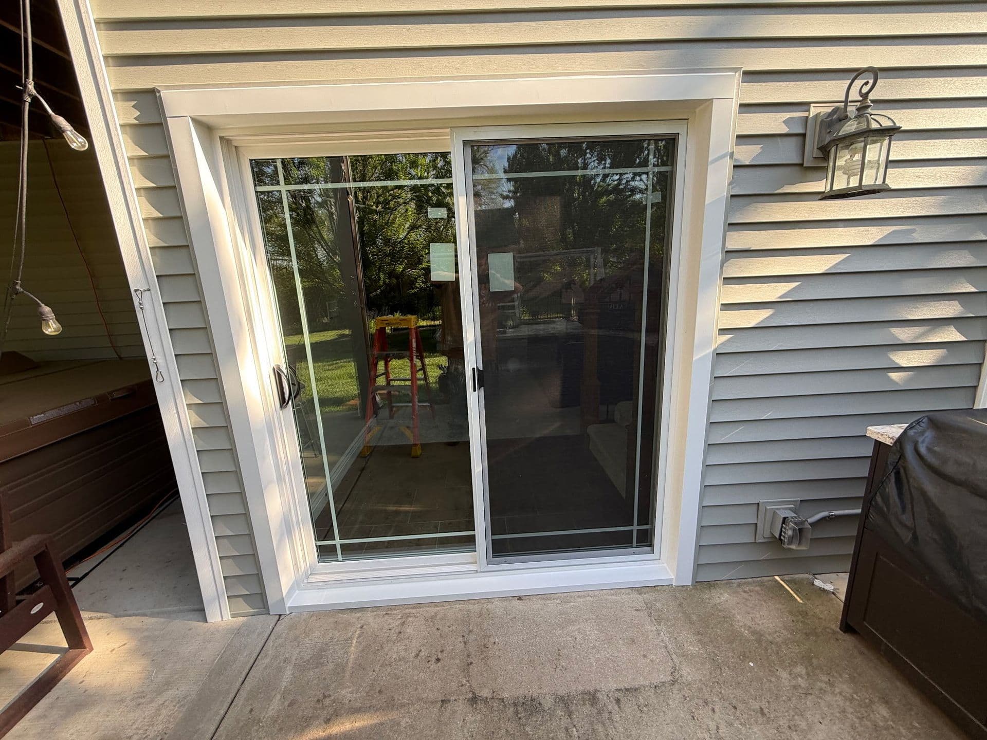 Sliding glass patio door in a gray-sided house, reflecting trees and lawn, with a grill, wall lantern, and concrete patio outside.