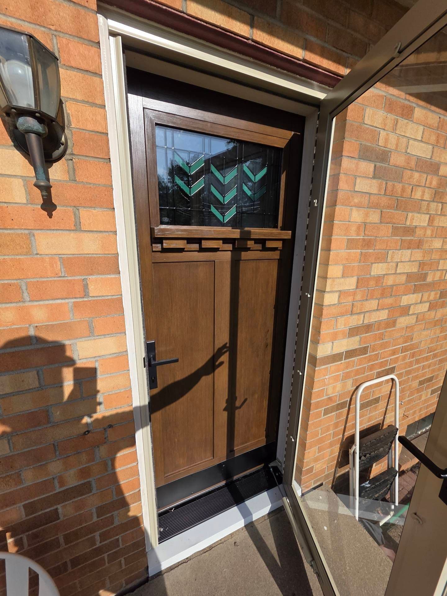 Sunlit brick entryway with a dark wooden front door, teal geometric glass panels, black handle, outdoor lamp, and leaning hand truck.