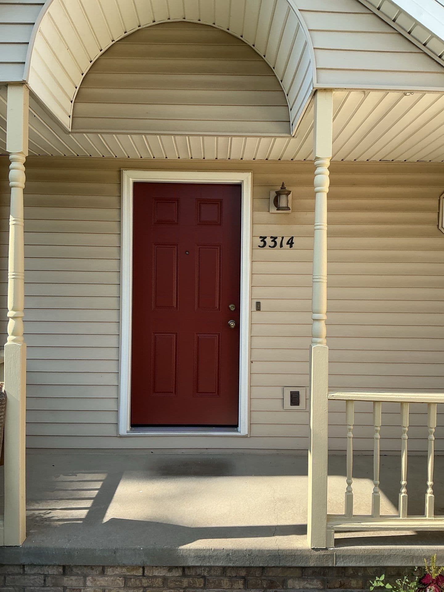 Front porch of a beige-sided home with a dark red door marked 3314, white columns, lantern, railing, and warm afternoon shadows.
