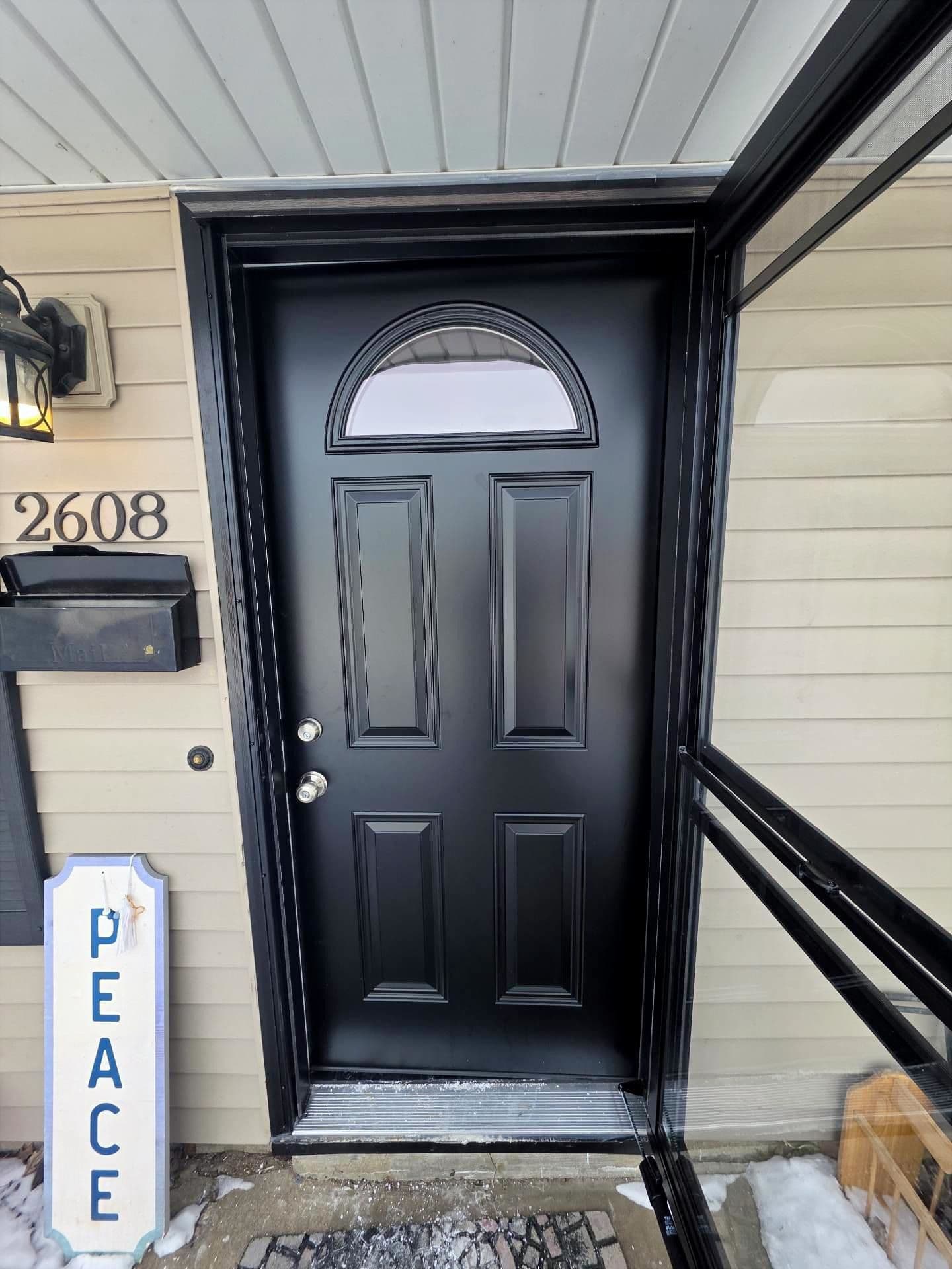 Black paneled front door with arched window, house number 2608, “PEACE” sign, snow, and porch details create a calm, welcoming entry.