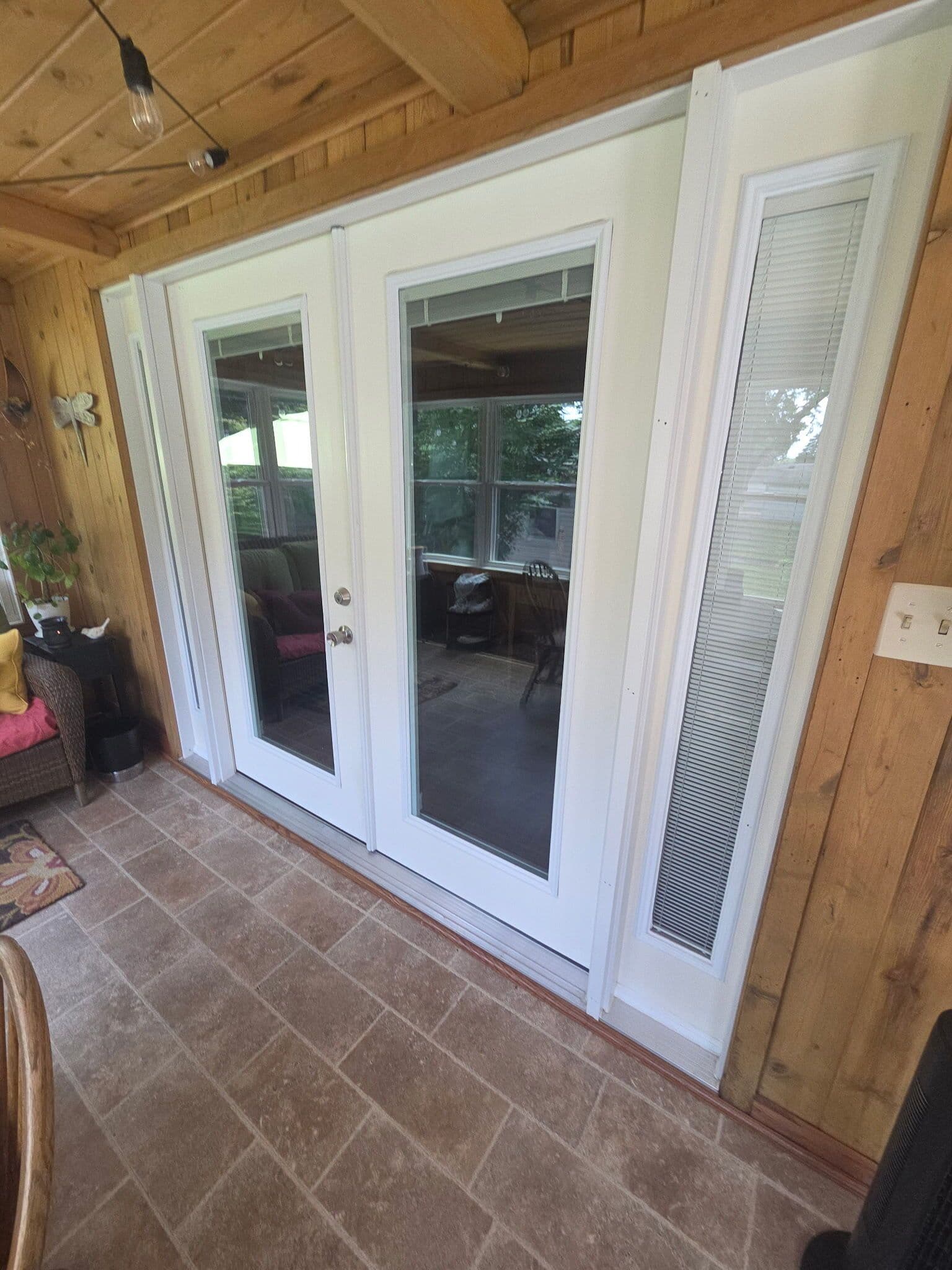 Wood-paneled sunroom with white French doors and sidelights, tiled floor, and windows overlooking greenery; bright, welcoming interior.