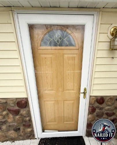 Closed light wood front door with decorative half-moon window, white trim, beige siding, stone accents, and “Nailed It” logo.